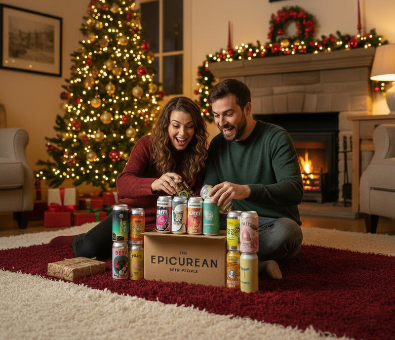 Man and woman opening a box of 'The Epicurean' beer cans in a festively decorated living room with Christmas tree and fireplace.